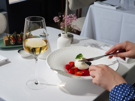 Une main sert un plat de tomates cerises et mozzarella, avec un verre de vin blanc sur une table élégante.